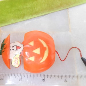 May include: Orange plastic Halloween toy shaped like a pumpkin with a ghost emerging from it. The pumpkin has a jack-o'-lantern face and a red string attached to a black cone. The ghost has a smiling face and the word "Boo!" on it.
