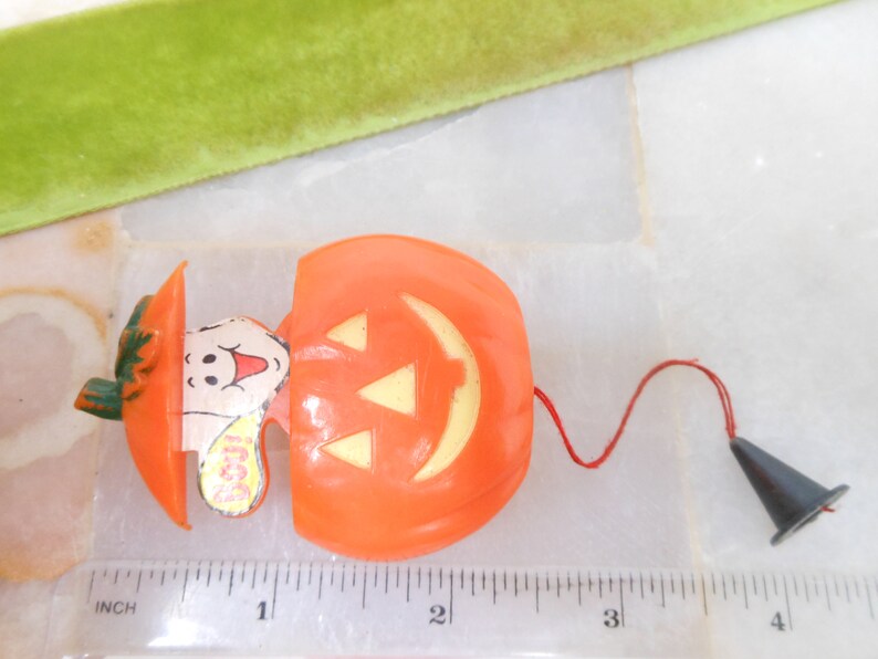 May include: Orange plastic Halloween toy shaped like a pumpkin with a ghost emerging from it. The pumpkin has a jack-o'-lantern face and a red string attached to a black cone. The ghost has a smiling face and the word "Boo!" on it.