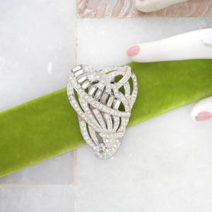 May include: A silver-toned, Art Deco-style brooch with a heart-shaped design, encrusted with sparkling rhinestones. The brooch is set against a vibrant green velvet ribbon, with a white marble surface in the background.