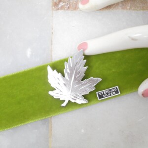 May include: A silver maple leaf pin on a bright green velvet ribbon. The pin is detailed with leaf veins and has a small rectangular tag that reads "STERLING SILVER". The ribbon is set against a white and tan background.