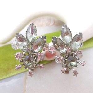 May include: A pair of silver tone clip-on earrings with a floral design. The earrings feature clear and light green rhinestones.
