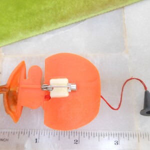 May include: Orange plastic pin with a white clasp and a small black cone attached by a red string. The pin is shaped like a pumpkin. A green velvet ribbon is in the background. The pin measures approximately 1 inch in length.