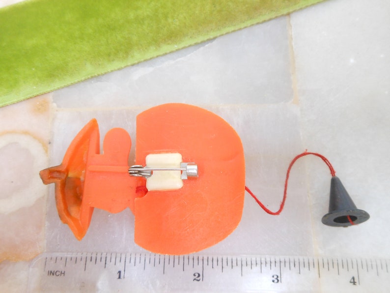 May include: Orange plastic pin with a white clasp and a small black cone attached by a red string. The pin is shaped like a pumpkin. A green velvet ribbon is in the background. The pin measures approximately 1 inch in length.
