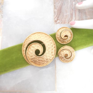 May include: A gold-toned jewelry set consisting of a circular brooch and a pair of matching round clip-on earrings. Each piece features a textured, woven-look surface with a prominent spiral design that curves inward toward the center.