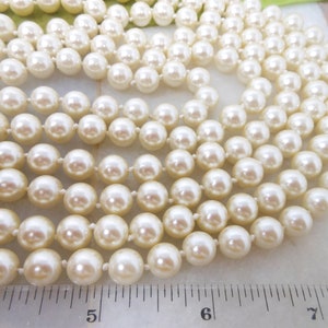 May include: A strand of white pearl beads, laid out on a white surface with a ruler for scale. The pearls are round and smooth, and they are strung on a thin, white thread.