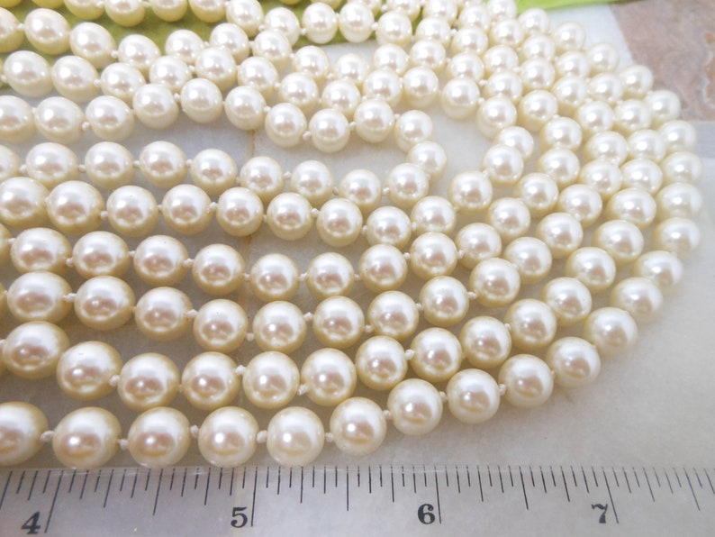May include: A strand of white pearl beads, laid out on a white surface with a ruler for scale. The pearls are round and smooth, and they are strung on a thin, white thread.