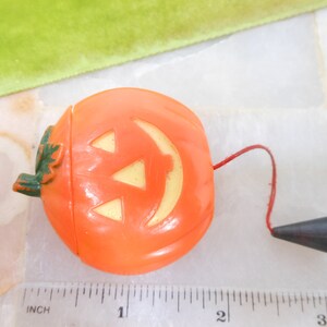 May include: Orange plastic Halloween jack-o'-lantern with a smiling face and a green leaf detail. It has a red string attached to a black cone-shaped piece. A ruler with inch measurements is in the foreground.