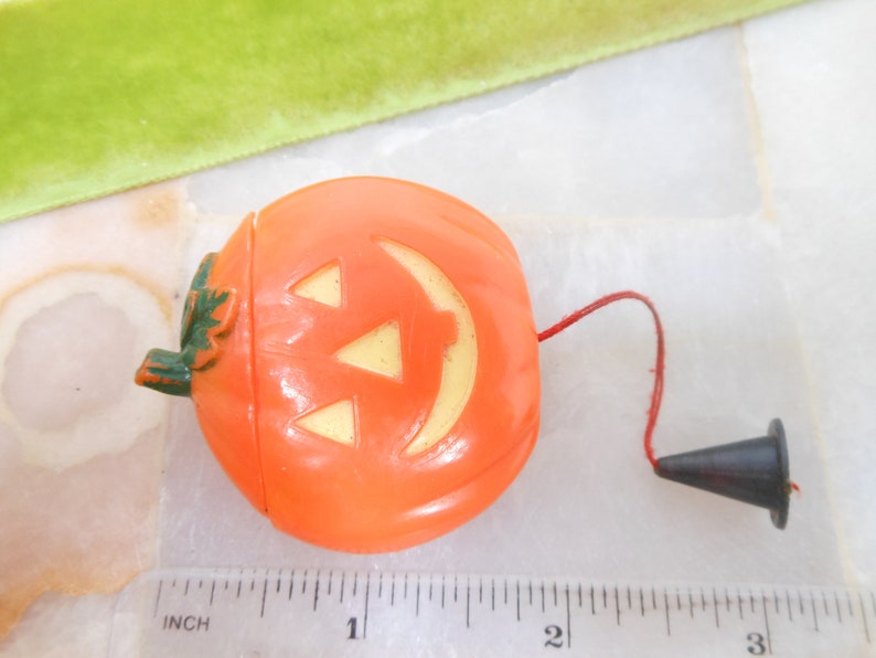 May include: Orange plastic Halloween jack-o'-lantern with a smiling face and a green leaf detail. It has a red string attached to a black cone-shaped piece. A ruler with inch measurements is in the foreground.