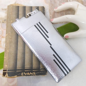 May include: A silver cigarette case with a hammered finish and black geometric design. The case has a hinged lid and a small, silver lighter attached to the top.