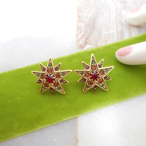 May include: A pair of star-shaped stud earrings with gold-tone metal and colorful gemstones. Each earring features a central red gemstone surrounded by smaller gems in shades of green, orange, and purple. The earrings rest on a green velvet ribbon.