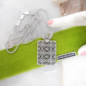 May include: A sterling silver necklace with a rectangular pendant featuring an openwork floral design. The pendant hangs from a delicate chain. The necklace rests on a green velvet surface, with a "STERLING SILVER" tag.