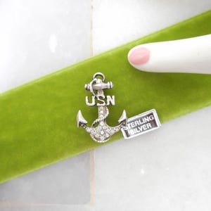 May include: A silver anchor charm with the letters "USN" and small clear stones. A small rectangular tag reads "STERLING SILVER". The charm rests on a bright green velvet ribbon.