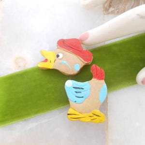 May include: Two wooden painted duck figurines. One duck is wearing a red hat and the other has blue wings. Both ducks have yellow beaks and brown bodies.
