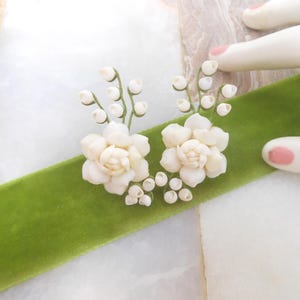 May include: Two white floral embellishments with delicate stems and buds are arranged on a vibrant green velvet ribbon. The ribbon is set against a soft, textured background. The image is a close-up shot.