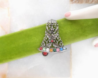 May include: A silver-toned bell-shaped brooch with intricate filigree detailing. The brooch is adorned with a row of colorful gemstones and a single red gemstone at the bottom. It rests on a vibrant green velvet ribbon.