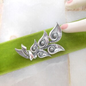May include: A silver-toned brooch with a leaf and swirl design, featuring small, round, gray accents. The brooch is set against a vibrant green velvet ribbon. The design is intricate and detailed.