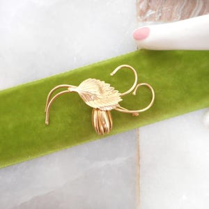 May include: A gold-toned floral brooch with a leaf and curved stem design. The brooch is set on a vibrant green velvet ribbon. The background is a marble surface.