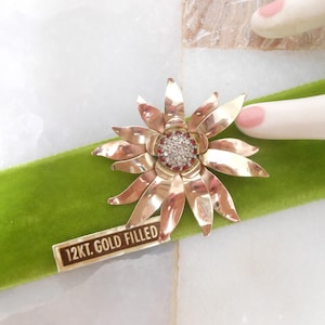 May include: A gold-toned flower brooch with a central cluster of clear and red rhinestones. The brooch has multiple petals and is labeled "12kt. Gold Filled". The brooch rests on a green velvet surface.