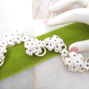 May include: A white floral necklace with small, detailed flowers and a cream-colored ring base. The necklace rests on a vibrant green velvet ribbon. The background is a marble surface.