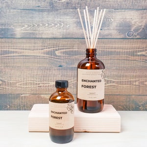 May include: Two amber glass bottles with brown labels that read "Enchanted Forest" reed diffuser. The larger bottle has a set of wooden reeds inserted into the top. The bottles are sitting on a light brown wooden surface.