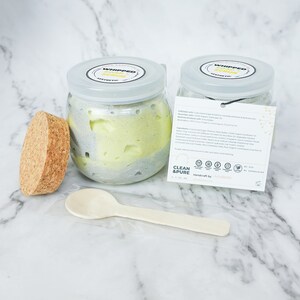 May include: A glass jar of whipped soap scrub with a gray and green swirl. The jar has a cork lid and a wooden spoon. The label on the jar reads "Whipped Soap Scrub" and includes a list of ingredients and instructions for use. The label also says "Clean & Pure" and "Handcraft by Aesthetic".