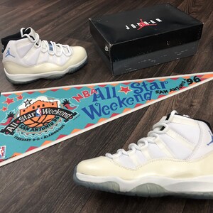 May include: A pair of white and blue Air Jordan 11 sneakers with a clear outsole. The sneakers are sitting on a wooden floor with a pennant that reads "NBA All Star Weekend San Antonio '96".