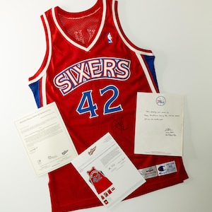 Puede incluir: Una camiseta de baloncesto roja con la palabra "SIXERS" y el número "42" en blanco y azul. La camiseta tiene ribetes blancos y azules. Varios documentos se colocan alrededor de la camiseta, incluida una carta de autenticidad.
