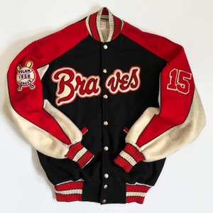 Puede incluir: Una chaqueta de béisbol negra, roja y blanca con "Braves" bordado en la parte delantera y "15" en la manga derecha. La chaqueta tiene un parche blanco en la manga izquierda con el texto "Polar 1958 League" y una pelota de béisbol y bates.