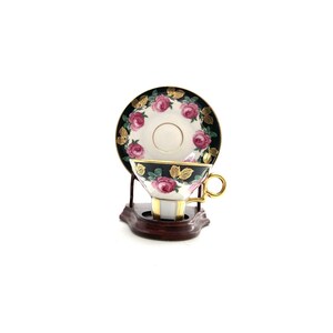 May include: A black and gold teacup and saucer set with pink roses and green leaves. The set is on a wooden stand.