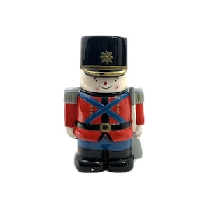 May include: A ceramic nutcracker figurine in the shape of a soldier. The soldier is wearing a red and blue uniform with a black hat and a gold belt. The nutcracker has a white face with black eyes and a red nose.