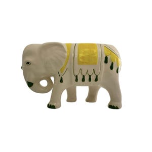 May include: White ceramic elephant figurine with a yellow and green saddle. The elephant has a trunk curled up and green details on its legs.
