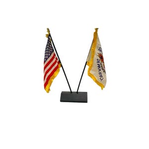 May include: Two small flags on a black base. One flag is the American flag with red and white stripes and blue stars. The other flag is white with a gold border and text. Both flags have gold fringe.