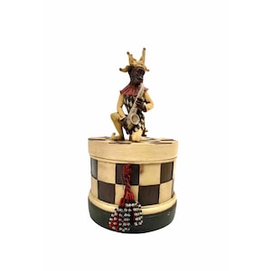 May include: A decorative round box with a checkerboard pattern in cream and dark brown. A figurine of a jester playing a saxophone sits atop the lid. The jester is dressed in a harlequin costume with a red and black color scheme. Red and black tassels hang from the box.