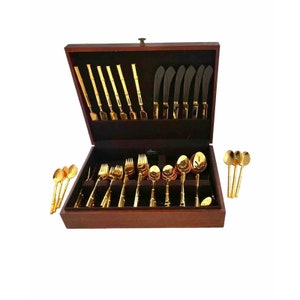 May include: A complete gold-colored flatware set displayed in a dark wooden box. The set includes spoons, forks, knives, and serving utensils. The utensils have a bamboo-style handle design.