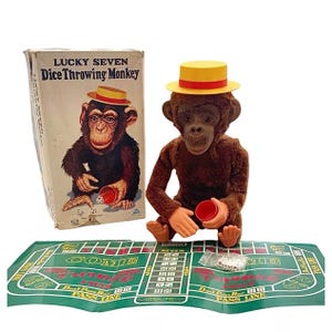 May include: A brown plush monkey wearing a yellow and red hat, holding a red cup and dice. The monkey is a vintage toy that throws dice. The box for the toy is in the background and says "Lucky Seven Dice Throwing Monkey".
