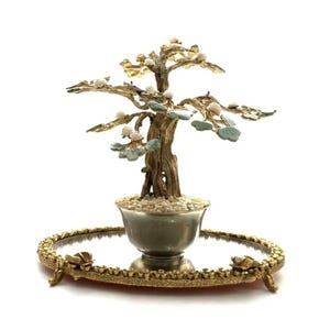 May include: A decorative bonsai tree sculpture with a detailed, textured trunk and delicate leaves. The tree sits in a silver-toned pot filled with small stones, placed on a gold-colored, ornate tray with a mirrored surface.