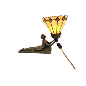 May include: A bronze sculpture of a woman holding up a stained glass lampshade. The lampshade is a light brown color with a geometric pattern and is lit from within.