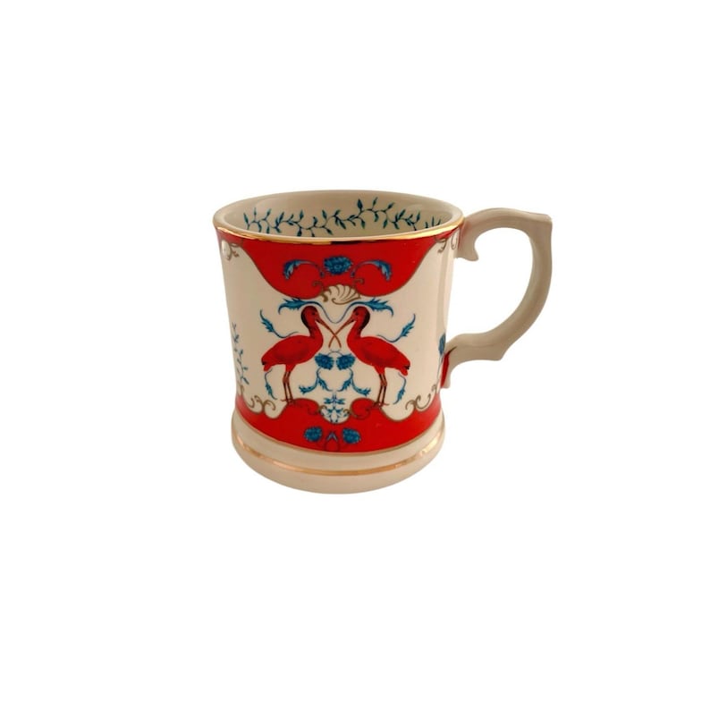 K&ouml;nnte beinhalten: Cremefarbene Keramiktasse mit einem roten und blauen Design mit zwei V&ouml;geln. Die Tasse hat einen goldenen Rand und Henkel. Das Design umfasst florale und scrollende Elemente. Ein dekoratives St&uuml;ck f&uuml;r die K&uuml;che oder die Inneneinrichtung.