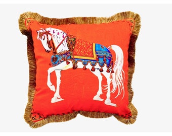 May include: Orange throw pillow with a white horse wearing a colorful saddle and bridle. The pillow has a gold fringe border.