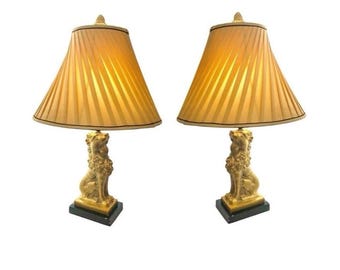 May include: Pair of gold-toned table lamps. Each lamp features a pleated, gold lampshade with a dark trim. The base of each lamp is a gold-colored lion statue on a black square base. The lamps are set against a white background.