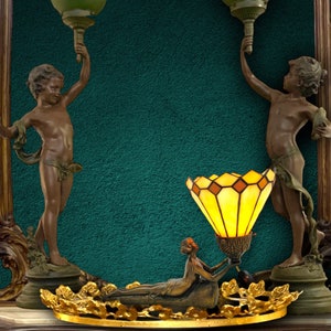 May include: A decorative lamp with a stained glass shade and a bronze base. The lamp is flanked by two cherub statues holding up green glass globes.