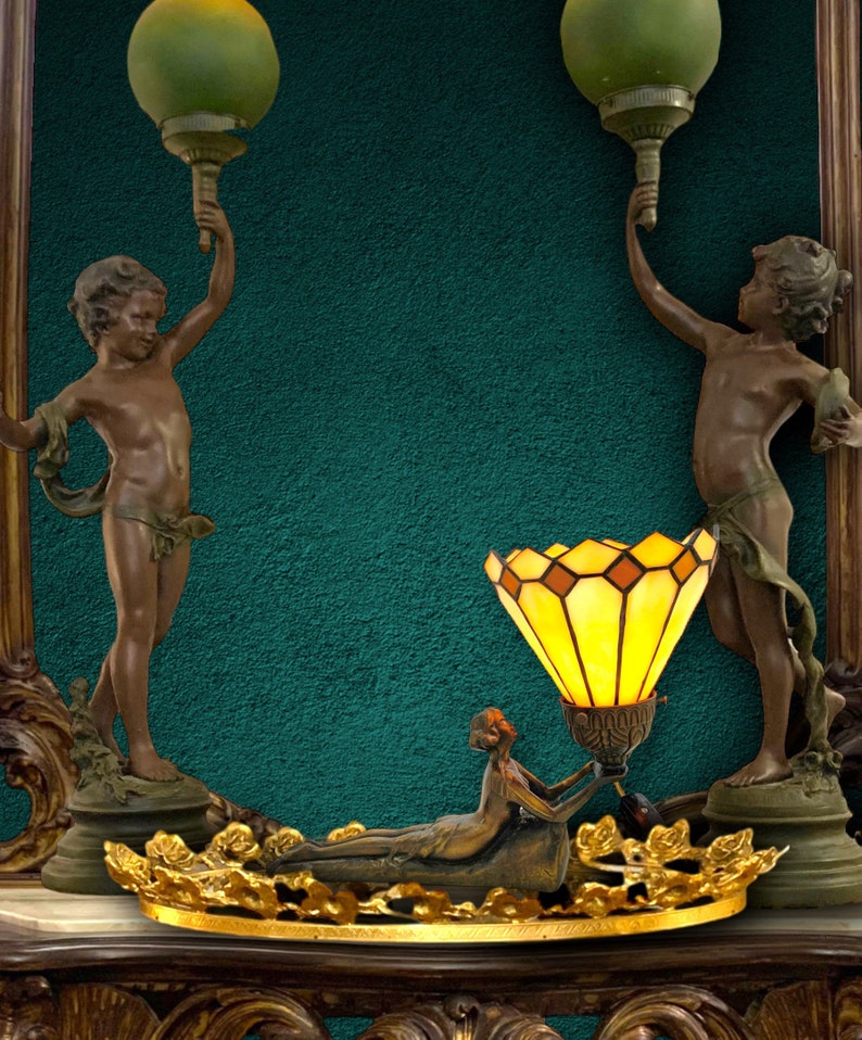 May include: A decorative lamp with a stained glass shade and a bronze base. The lamp is flanked by two cherub statues holding up green glass globes.