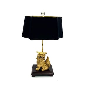 May include: A gold-painted Foo dog table lamp with a black lampshade. The lamp base is a square wooden platform.