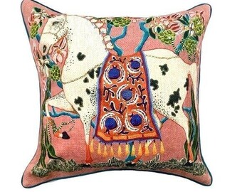 May include: Decorative square pillow with a pink background and a detailed illustration of a white horse with black spots. The horse is adorned with a patterned orange and blue blanket. The pillow has a dark blue border and features floral and vine accents.