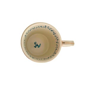 K&ouml;nnte beinhalten: Cremefarbene Keramik-Tasse mit goldenem Rand und Griff. Der Innenraum hat ein t&uuml;rkisfarbenes Blumendesign am Rand und einen t&uuml;rkisfarbenen floralen Akzent am Boden. Der Griff hat einen goldenen Streifen.