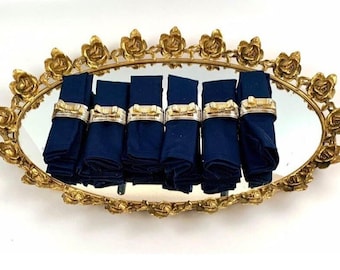 May include: An oval tray with a mirrored surface and a decorative rose border, finished in gold. Six navy blue napkins are arranged on the tray, each with a gold and silver napkin ring.