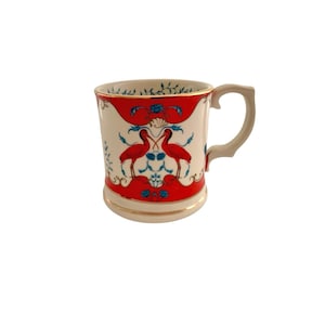 May include: A ceramic mug with a cream-colored base and handle, featuring a red band with a design of two red birds and blue floral accents. The rim and base have gold trim. The mug is designed for beverages.