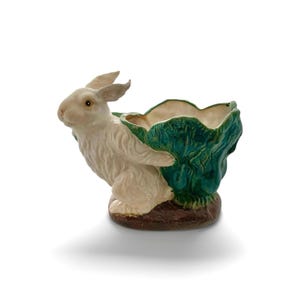 May include: A ceramic planter featuring a white rabbit holding a green cabbage-shaped bowl. The rabbit has detailed fur and a brown base. The bowl has a textured, leafy design.