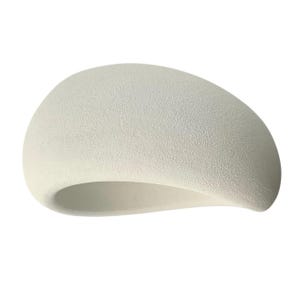 May include: A cream-colored, abstract sculpture with a textured surface. The form is a smooth, curved shape with a hollowed-out interior, resembling a stylized beret or a smooth, organic form.