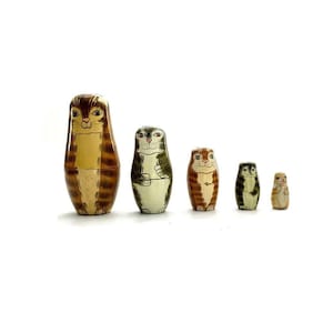 May include: Set of five nesting dolls, each depicting a cat with different colored fur. The largest doll is brown and yellow, the next is green and white, the next is orange and white, the next is gray and white, and the smallest is white with brown spots.